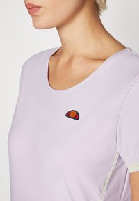 Lyselilla kortermet topp med rund hals og et brodert logo i oransje og rødt på brystet. Har subtile teksturerte stoff og kontraststing.