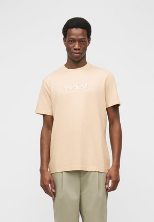 Mladý muž stojaci oblečený v béžovom tričku s logom GUESS a svetlosivých nohaviciach na jednoduchom svetlosivom pozadí.