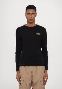 Camisola de manga longa preta feita de algodão. Apresenta um pequeno logótipo dourado no peito, decote redondo e um design ajustado.