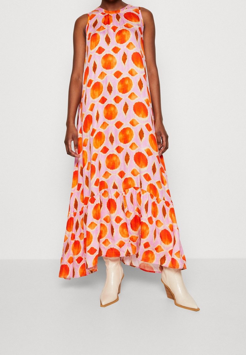 Femme portant une robe longue fluide sans manches avec des motifs circulaires orange et rouge et des bottines blanches à talons en bois.