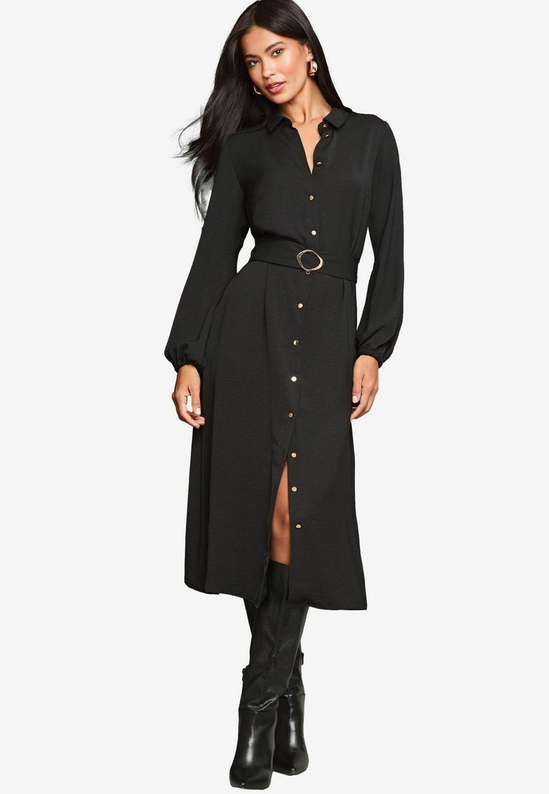 Schwarzes Langarmkleid mit Knopfleiste und tailliertem Gürtel. Verfügt über goldene Knöpfe und einen seitlichen Schlitz. Das Modell kombiniert es mit kniehohen Stiefeln.