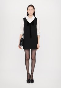 Zwarte mouwloze jurk met een fluwelen strik, gedragen over een wit blouse met kraag. Inclusief zwarte doorzichtige panty's en zwarte chunky schoenen.