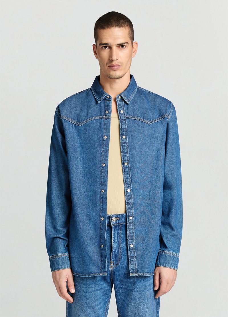 Jeanshemd mit Knopfleiste vorne, hellblauer Farbe, langen Ärmeln und kontrastierenden Nähten. Wird über einem beigen Unterhemd getragen.