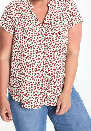 Mujer con una blusa blanca de manga corta con patrones de cerezas rojas y hojas verdes combinada con jeans azules contra un fondo liso.