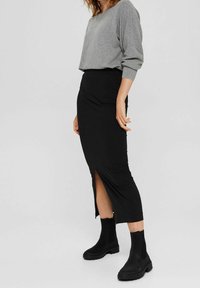 Haut gris à manches longues avec poignets côtelés, associé à une jupe moulante noire mi-genou avec une fente latérale, portée avec des bottines noires.