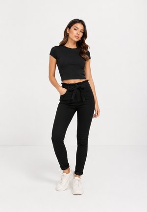 Frau trägt ein schwarzes Crop-Top, hoch taillierte schwarze Hose mit gebundenem Gürtel und weißen Sneakers, steht mit einer Hand in der Tasche.