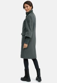 Manteau en laine mélangée vert foncé avec un col haut, des poches latérales et des manches larges, porté avec des bottes noires à la cheville et un pantalon noir ajusté.