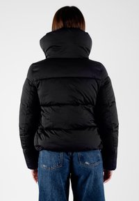 Giubbotto imbottito nero con colletto alto, texture trapuntata e polsini a coste, abbinato a jeans in denim blu dalla vestibilità ampia.