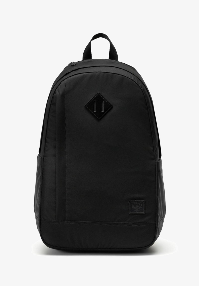 Sac à dos en tissu noir avec poignée supérieure, forme arrondie, pièce en cuir en forme de losange à l'avant et petite étiquette de marque rectangulaire près du bas.