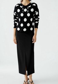 Un pull noir à pois blancs avec une capuche, associé à une jupe midi noire avec une fente sur le devant, porté avec des sandales noires.