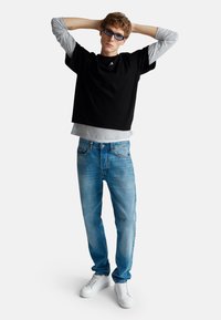 Mladý muž v brýlích na slunce má na sobě černou košili s krátkým rukávem přes šedé tričko s dlouhým rukávem, modré džíny a bílé tenisky, stojí s rukama za hlavou.