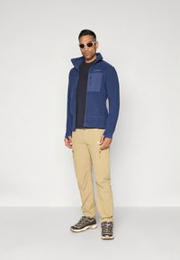 Temno modra flis jakna z visokim ovratnikom in žepom na prsih z zadrgo, kombinirana s ten barvnim hlačami in večbarvnimi pohodniškimi čevlji. Bele sončna očala.