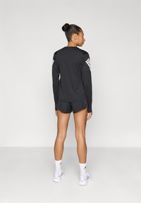 Čierny športový dlhý rukáv s bielymi pruhmi na ramenách, kombinovaný s čiernymi krátkymi nohavicami. Modelka má na sebe biele ponožky a sivé topánky s ružovými akcentmi.