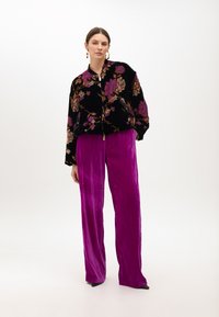 Zwarte fluwelen bomberjack met bloemmotieven, gecombineerd met paarse satijnen, plissé hoge taille broek. Subtiele metalen accenten aanwezig.