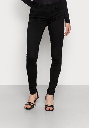 Femme portant un jean skinny noir et des sandales à talons noires à lanières, debout devant un fond clair uni.