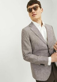 Blazer gris claro texturizado con un cierre de un solo botón, solapas con muesca y dos botones negros en el puño. Llevado sobre una camisa blanca.