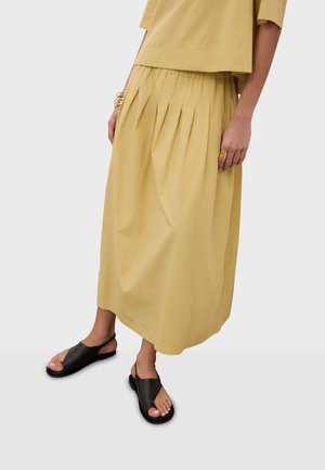 Femme portant une jupe midi plissée jaune et un haut, accessoirisée avec des bagues et bracelets en or, ainsi que des sandales plates noires à bout ouvert.