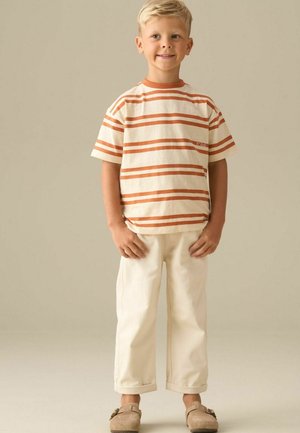 Ung dreng står iført beige bukser, en hvid og orange stribet t-shirt og lysebrune slip-on sko, mod en ensfarvet beige baggrund.