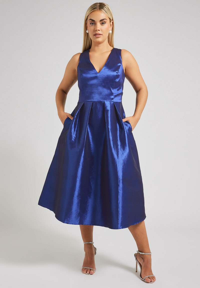 Sininen satiinimekko, jossa on V-kaula-aukko, olkaimeton malli ja A-linjainen hame, jossa on taskut ja sileä, kiiltävä pinta.