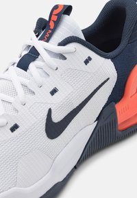 Białe buty sportowe z teksturowaną siateczką, granatowymi akcentami, pomarańczowym piętkiem i czarnym logo. Posiadają białe sznurówki i niebieską pętlę.