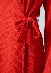 Vestido rojo texturizado con diseño envolvente, adornado con un lazo atado en la cintura. La tela presenta un patrón acanalado para mayor interés visual.