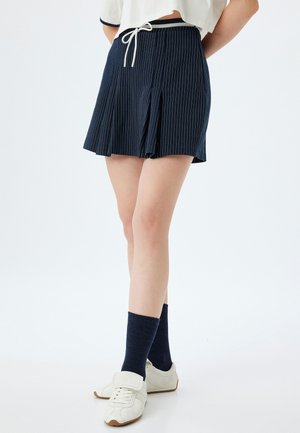 Marineblauwe geplooide rok met witte streepjes, elastische tailleband en trekkoord. Draag het met marineblauwe sokken en witte sneakers.