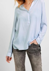 Femme portant un chemisier à rayures verticales bleu clair et blanc avec des manches longues et deux boutons, assorti à un jean noir délavé.