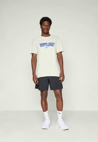 Lys grå t-shirt med blå tekst "SEJR STARTER MED TRÆNING" og logo, parret med sorte shorts og hvide sneakers.