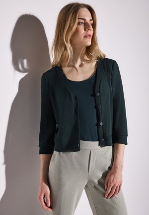 Cardigan texturé vert foncé à manches trois-quarts, avec un devant boutonné, associé à un pantalon taille haute gris clair.