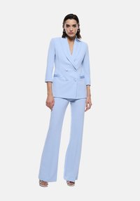 Completo su misura blu chiaro con giacca doppiopetto e pantaloni svasati. Presenta un collo a revers e sottili strisce a contrasto. Tessuto liscio.