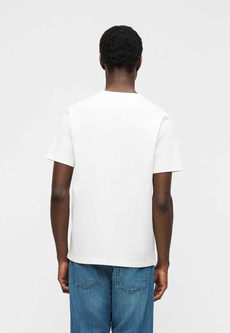 Personne aux cheveux courts et bouclés portant un t-shirt blanc uni à manches courtes et un jean bleu, debout de dos devant un fond neutre.