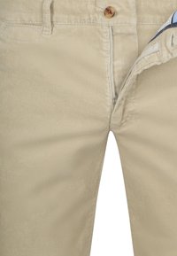 Beige ribfluwelen broek met gedeeltelijk open ritssluiting en bruine knoop in de tailleband, waardoor de gestreepte binnenstof zichtbaar is.