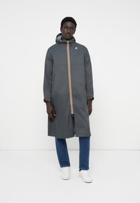 Halljas hall pikk mantel kapuutsiga, millel on kollane ja oranž vertikaalne triip ning kontrastsed detailid. Mudel kannab teksaseid ja valgeid sneaksereid.