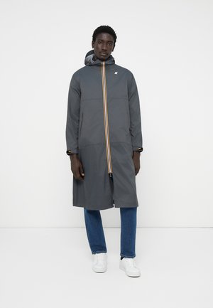 Γκρίζο μακρύ παλτό με κουκούλα, που διαθέτει κατακόρυφη κίτρινη και πορτοκαλί ρίγα και αντίθετες λεπτομέρειες από μέταλλο. Το μοντέλο φοράει τζιν και λευκά sneakers.