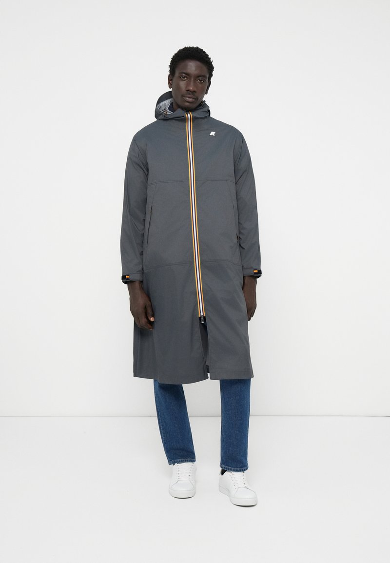 Halljas hall pikk mantel kapuutsiga, millel on kollane ja oranž vertikaalne triip ning kontrastsed detailid. Mudel kannab teksaseid ja valgeid sneaksereid.