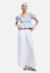 Blaues karriertes Smock-Crop-Top mit Puffärmeln, kombiniert mit einem weißen Maxikleid. Dazu flache blaue Sandalen mit Knotendetail.