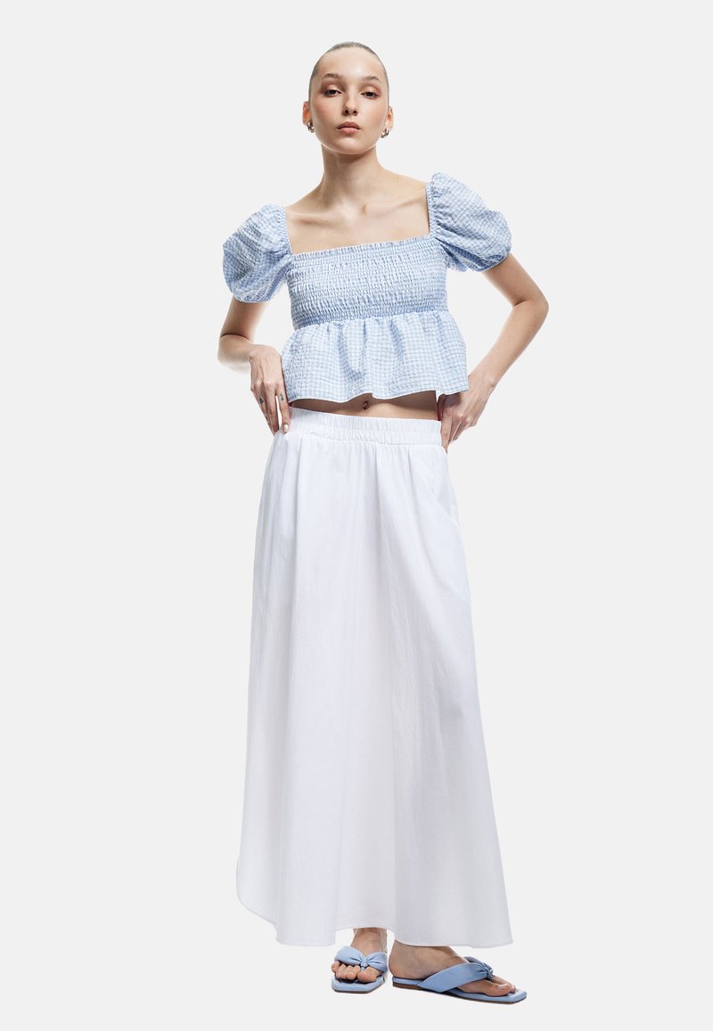 Blaues karriertes Smock-Crop-Top mit Puffärmeln, kombiniert mit einem weißen Maxikleid. Dazu flache blaue Sandalen mit Knotendetail.