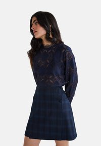 Haut à manches longues en dentelle bleu marine avec des motifs géométriques et un col festonné ; associé à une mini-jupe à carreaux bleu marine ornée de deux boutons.