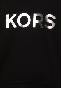 Černá tkanina s hladkým povrchem, na které je velkými metalickými stříbrnými písmeny napsáno "KORS". Design je jednoduchý a centrální.