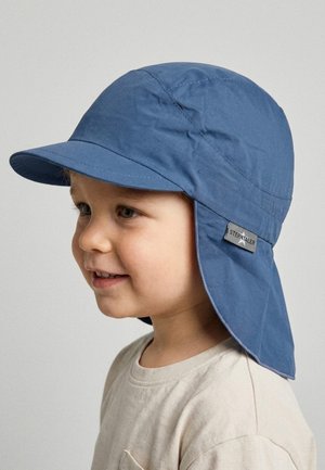 Toddler wearing beige shirt and blue sun hat with neck flap, smiling and looking left against a plain light background.