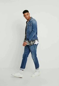 Casaco de ganga com lavagem azul clara, sobre uma camisa xadrez preto e branco. Jeans justos azuis e ténis brancos completam o look.