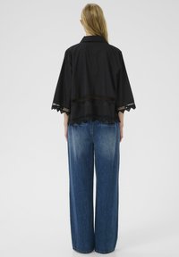 Sort skjortejakke med korte, brede ærmer og bølget kant. Parret med lyseblå, vide denimjeans. Minimalistisk design.
