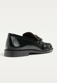 Schwarze Lederloafers mit glänzender Oberfläche, versehen mit einem geflochtenen Detail an der Vorderseite und einer niedrigen, flachen Gummisohle. Inklusive goldfarbener Akzentbeschläge.