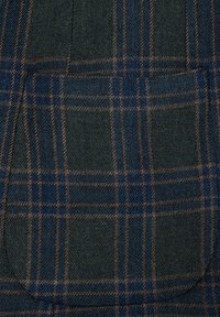 Wełniana tkanina w ciemnozielonym kolorze z niebiesko-brązowym wzorem w kratę; ma dwie przednie kieszenie o zaokrąglonych krawędziach oraz miękką teksturę.