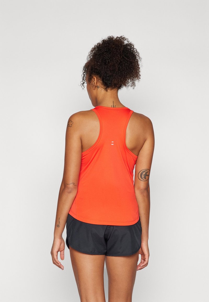 Vrouw met krullend haar in een oranje sporttop en zwarte shorts die met haar rug naar de camera staat tegen een effen achtergrond.