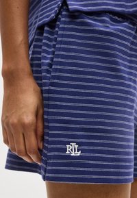 Χέρι που ακουμπά δίπλα σε μπλε και λευκή κοντή βερμούδα με λεπτές ρίγες και κεντημένο λογότυπο "RL" κοντά στο τελείωμα.