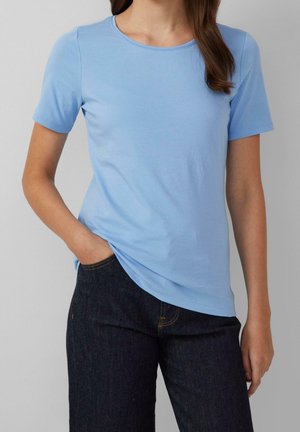 Kvinde iført en lyseblå kortærmet t-shirt og mørke jeans, står med den ene hånd i lommen foran en ensfarvet baggrund.