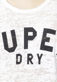 Witte katoenen t-shirt met zwarte, vetgedrukte tekst "SUPER DRY." De stof heeft een subtiel textuurpatroon. Ronde halslijn.