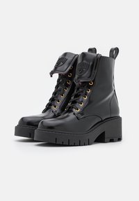 Schwarze Kampfstiefel aus lackiertem Leder mit einem klobigen Plateau-Absatz, goldenen Ösen und einer umgeschlagenen Lasche mit einem Designdetail.