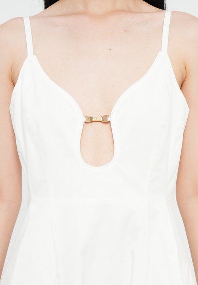 Vestido blanco con un escote en V profundo y tirantes finos, con un detalle de broche dorado en el escote y un corte en la parte trasera.